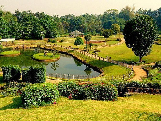A scenic view of the Pune-Okayama Friendship Garden, showing a winding path leading to a small bridge over a pond. Lush green lawns and trees surround the pond, creating a peaceful atmosphere.